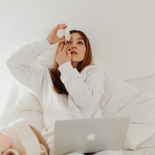 A woman wearing white sits in her bed applying Thealoz® Duo Eye Drops to her eyes