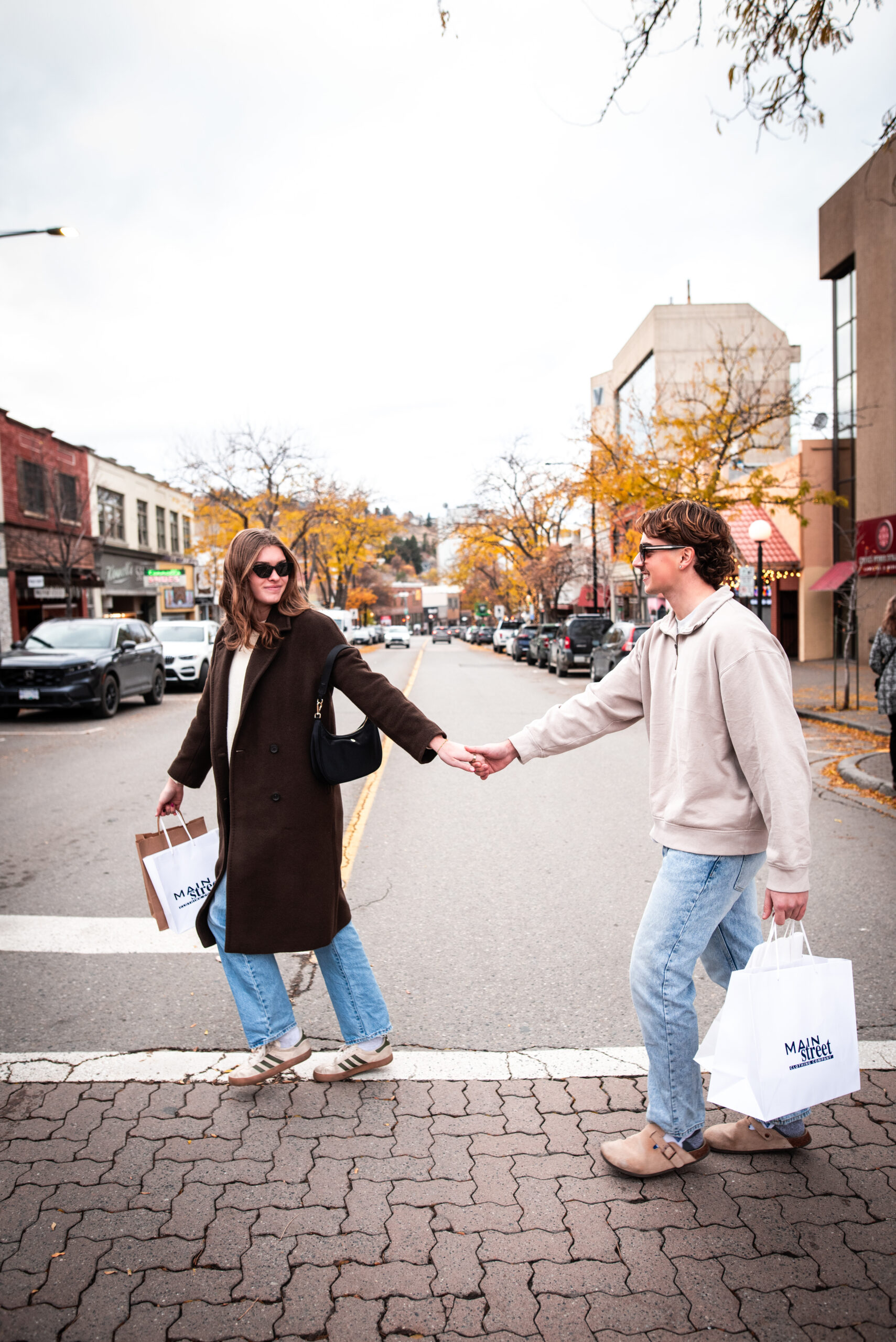 Two people wearing sunglasses hold hands while crossing a downtown street, carrying shopping bags and enjoying a fall day in the city.