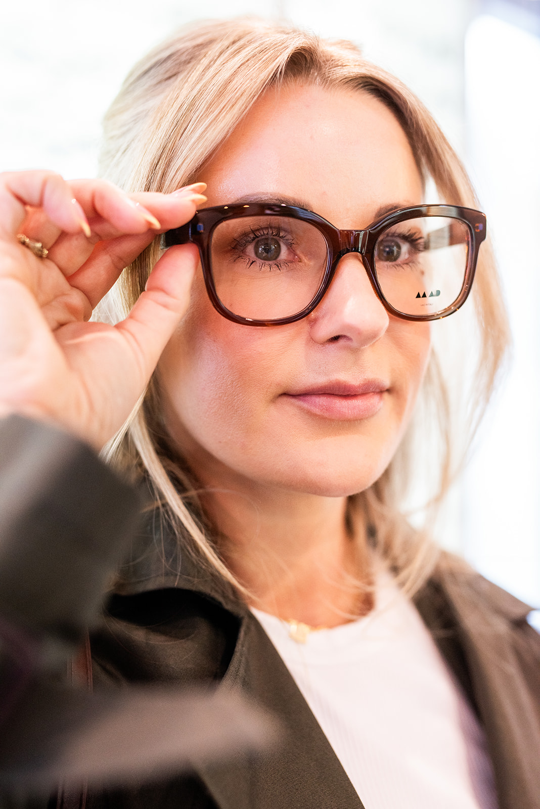 Woman trying on eyeglasses inside an optical boutique, standing in front of a wall display of frames.