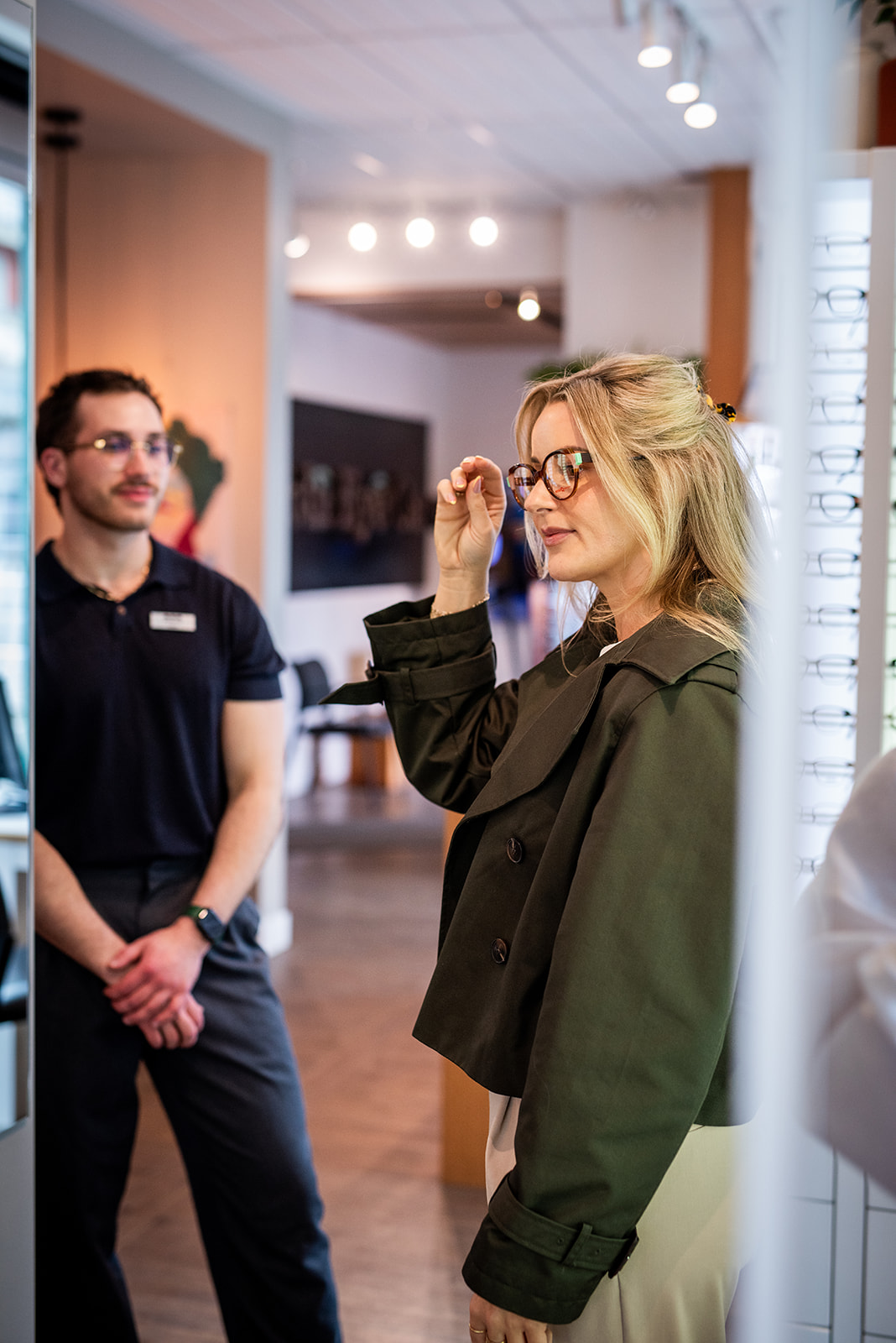 Woman trying on eyeglasses inside an optometry clinic while an optician stands nearby, offering assistance during a frame fitting.