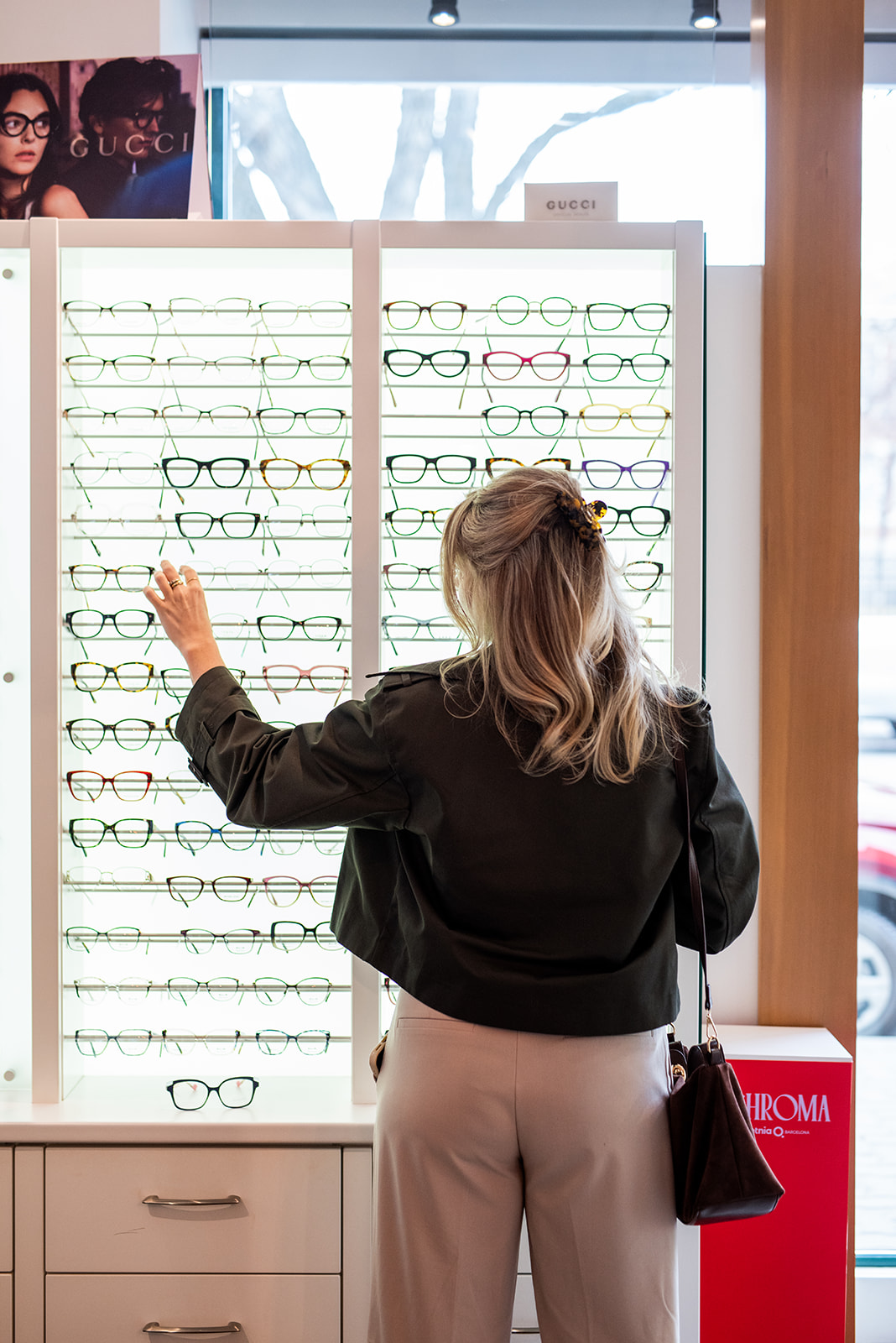 Woman browsing eyeglass frames inside an optometry clinic, reaching toward a wall display of glasses while standing in front of illuminated frame shelves.
