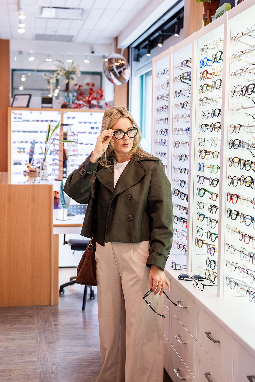 Woman browsing eyeglass frames inside an optometry clinic, holding a pair of glasses while viewing a large frame display.