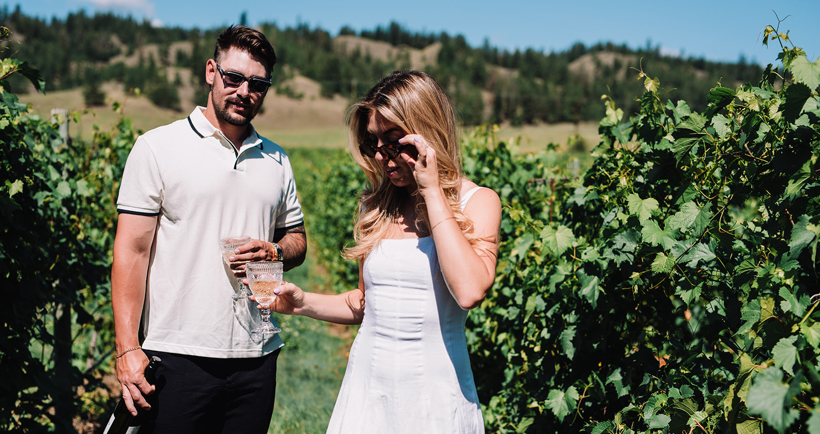 A man and woman standing outdoors in bright sunlight, wearing sunglasses and holding drinks while enjoying a sunny day among green vineyard rows.